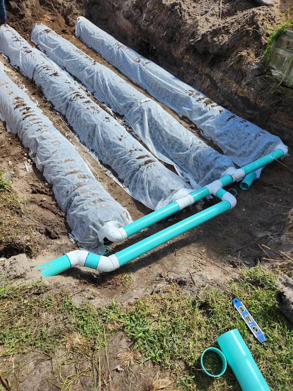 An excavated trench reveals four long, white fabric-wrapped drainage pipes partially covered with dirt. Bright turquoise PVC pipes with white fittings are laid in the foreground, connecting to the wrapped pipes, with a spirit level and pipe sections on the grassy ground.