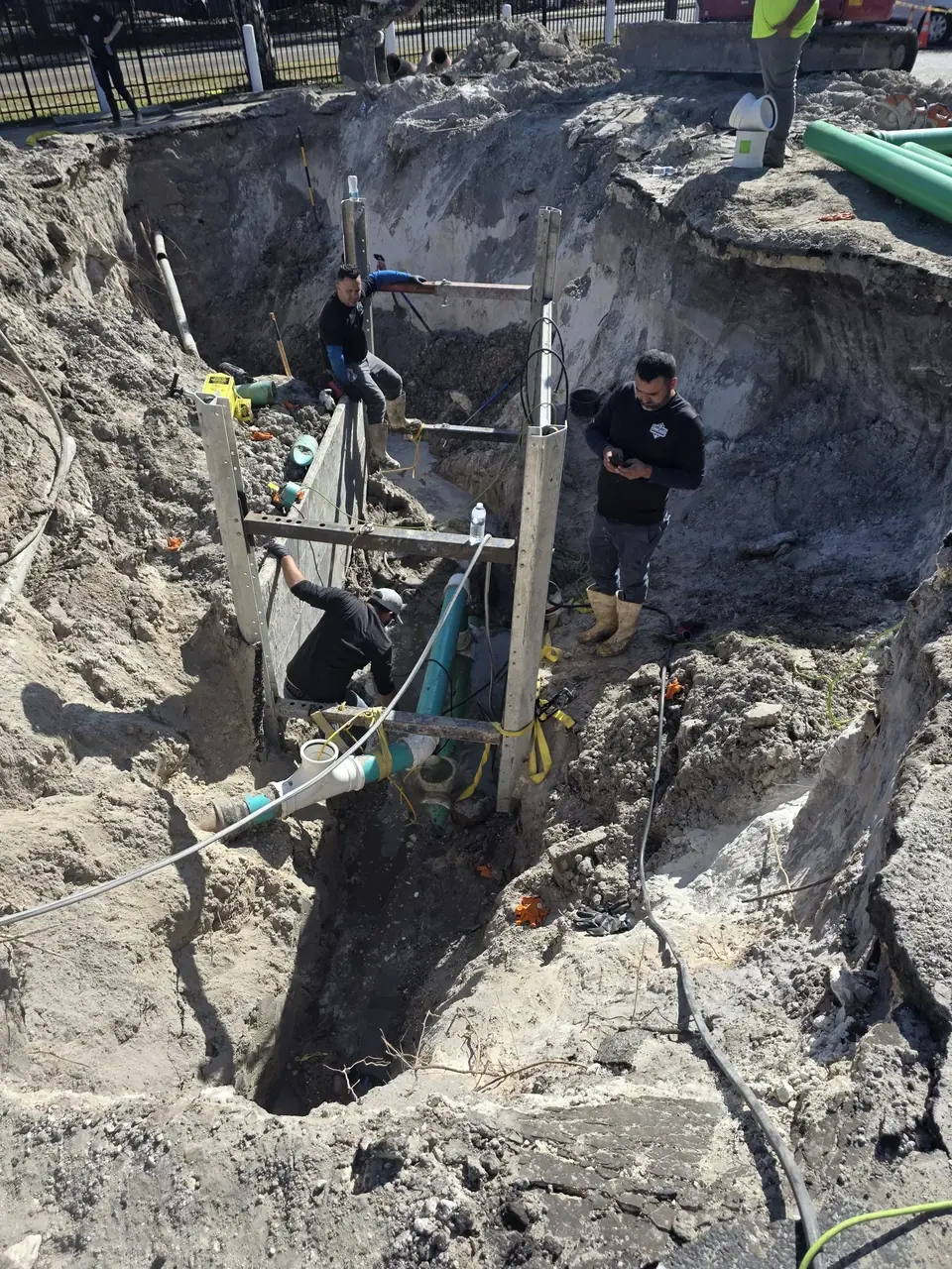 A large construction trench is dug into the ground, with several workers inside and around it. Green and white pipes are visible within the trench, supported by shoring.