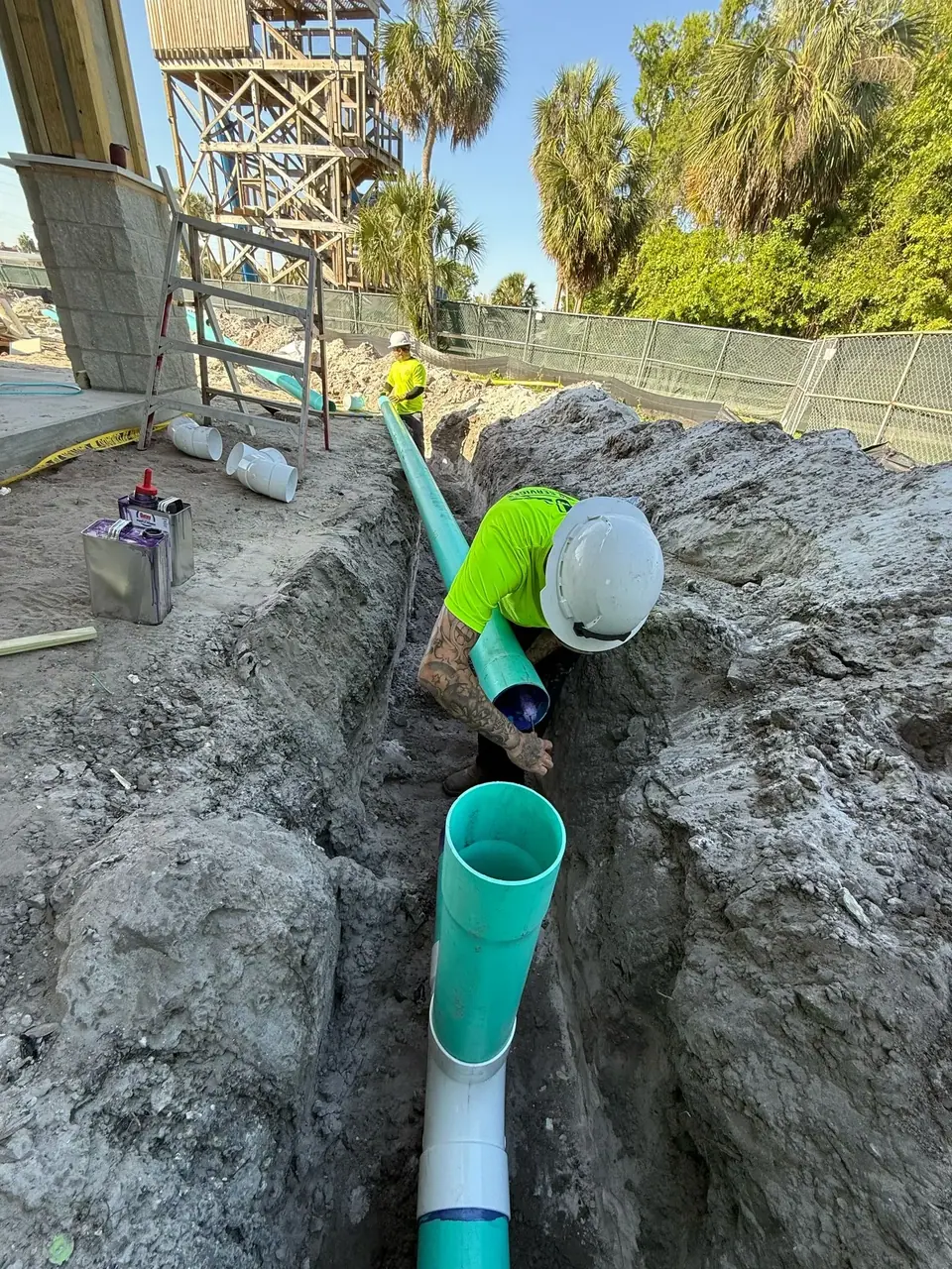 Two construction workers are installing green pipes in a dirt trench at a construction site. One worker in a hard hat and neon shirt is bent over in the trench, connecting pipe sections, with another worker standing nearby.