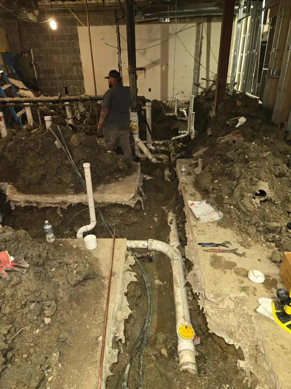 An indoor construction site with trenches dug into the dirt floor, revealing white PVC plumbing pipes. A worker in a dark shirt and cap stands in a trench, while another worker is visible on a ladder in the background.
