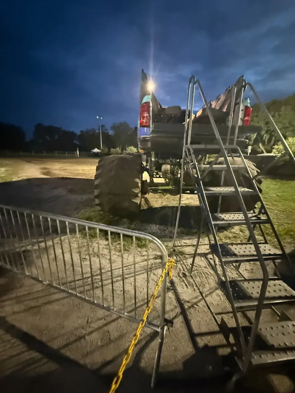 A monster truck is viewed from the rear at night, with a metal staircase providing access to its bed. A metal barrier with a yellow chain is in the foreground, separating the viewer from the truck.