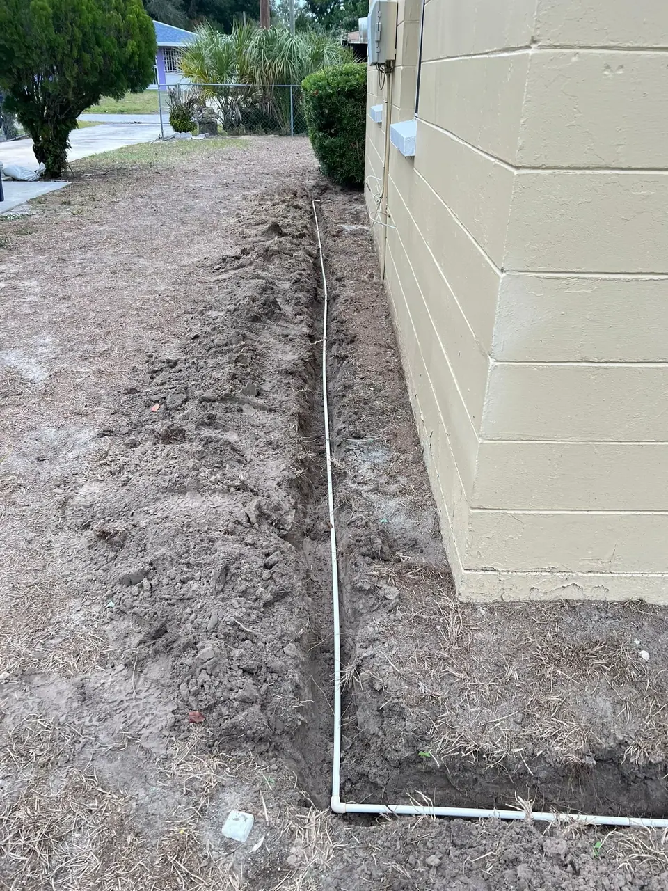 A narrow trench is dug in the dirt next to a light tan concrete block building, with a white pipe laid inside the trench and along the building's base. The surrounding ground is mostly bare dirt with some dry grass.