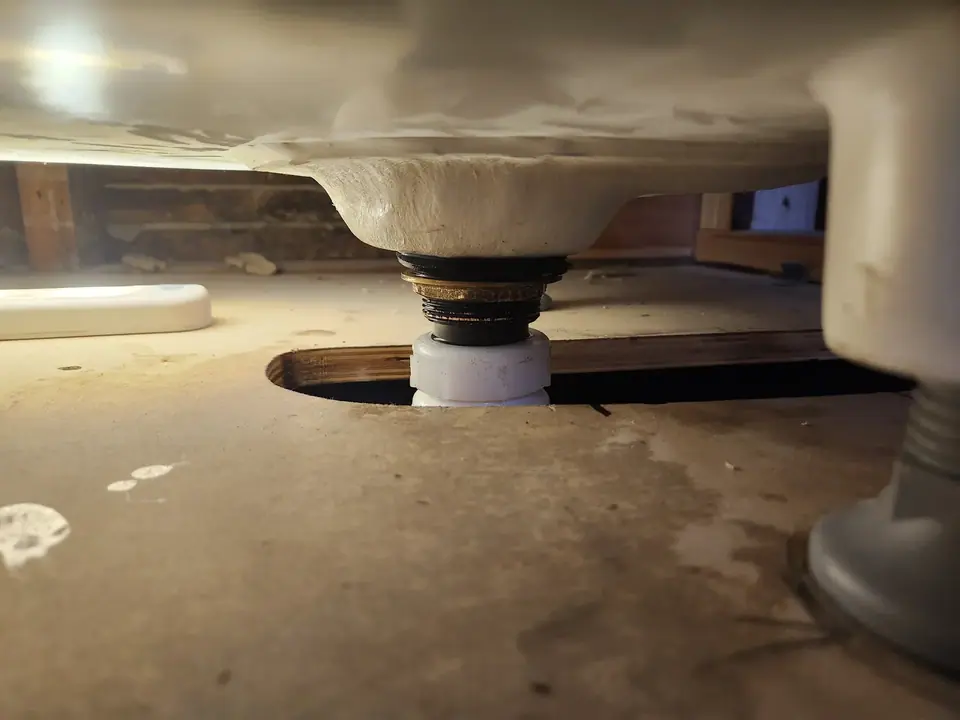 A low-angle view shows the underside of a white sink or toilet, with its drain assembly extending through a circular hole in a light brown wooden floor. The drain features brass-colored rings and connects to a white PVC pipe below the floor, with a gray pipe visible on the right.