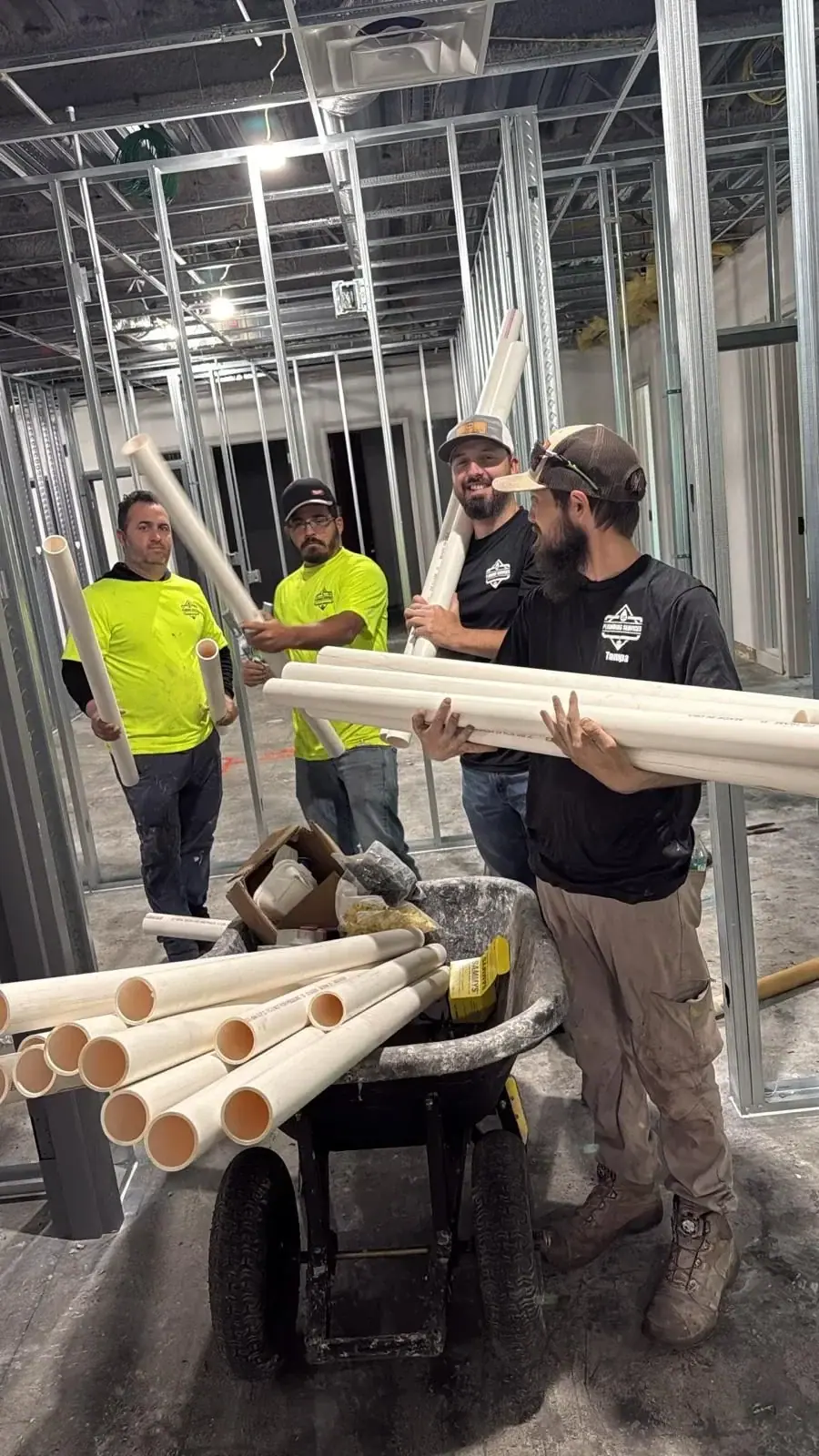 Four plumbers are standing in a room framed with metal studs, holding and organizing white PVC pipes. A wheelbarrow filled with pipes and other materials is in the foreground.