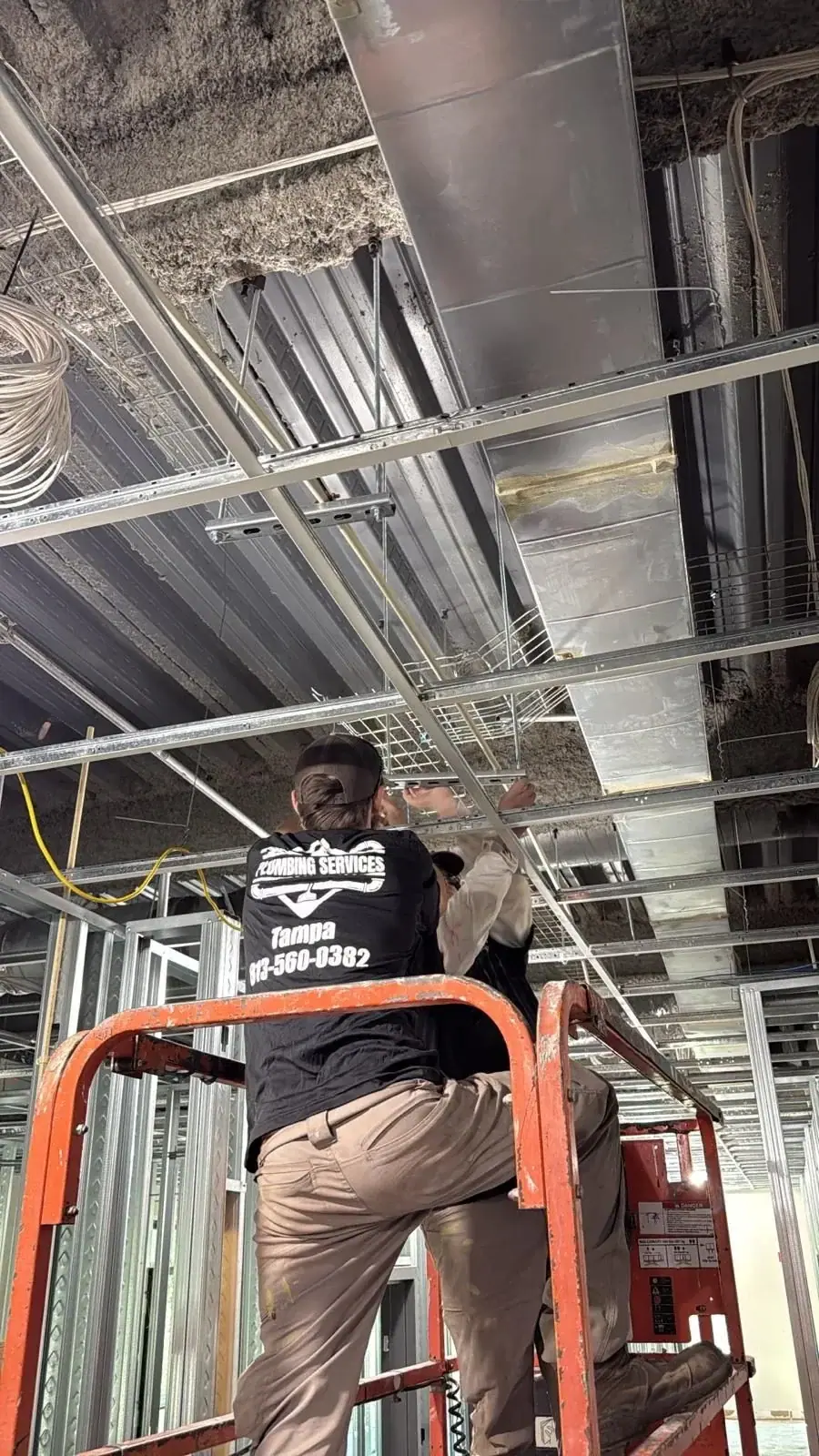 A plumbing technician, wearing a black shirt with "Plumbing Services Tampa" visible, stands on an orange scissor lift, working on overhead pipes and ductwork in a commercial building under construction. The ceiling shows exposed insulation, metal decking, and various conduits and structural elements.