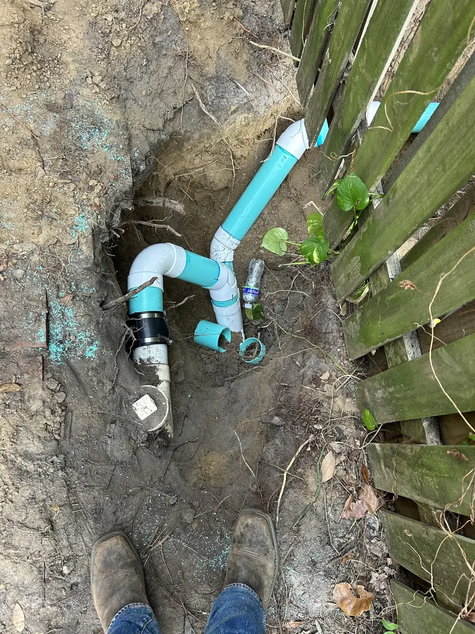 An overhead view shows a trench in the dirt with white and teal PVC plumbing pipes, some connected and some loose. A person's feet in brown work boots are visible at the bottom, next to a wooden fence on the right.