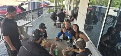 A group of people are gathered on an outdoor patio with picnic tables next to a building with large windows. In the foreground, a man makes a peace sign while a woman smiles at the camera, with portable toilets visible in the background.