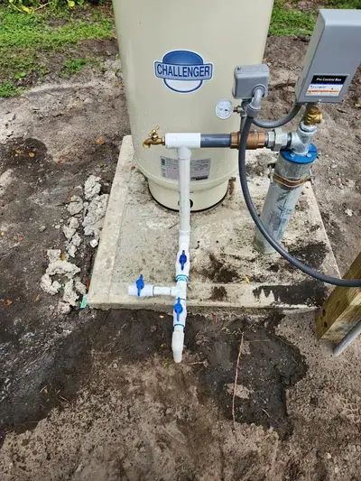 A "CHALLENGER" brand water pressure tank sits on a concrete pad, connected to a well pump system with PVC and metal pipes, blue-handled valves, a pressure gauge, and a "Pro Control Box." The system is installed outdoors on dirt ground with some foliage.