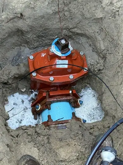 An orange water valve assembly is partially buried in a muddy pit, connected to a light blue pipe at its base. Water with white foam is pooling around the valve, and a black cable runs across its top.