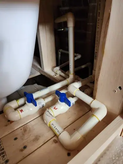 CPVC plumbing pipes with blue shut-off valves are installed on a wooden floor next to a white bathtub, with wooden framing and a brick wall visible in the background.
