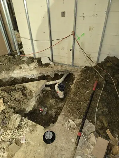 An overhead view of a construction site shows a trench with white PVC plumbing pipes installed in the dirt. A black shovel rests on the right, and unfinished walls with metal studs are visible in the background.