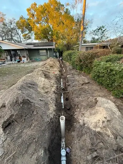 A long trench is dug in a grassy yard, with white PVC pipes laid along its length. Piles of excavated dirt line both sides of the trench, and a house with solar panels is visible in the background.