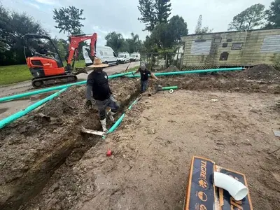 Two men are working in a muddy trench, installing green pipes, with an excavator and a mobile home visible in the background.