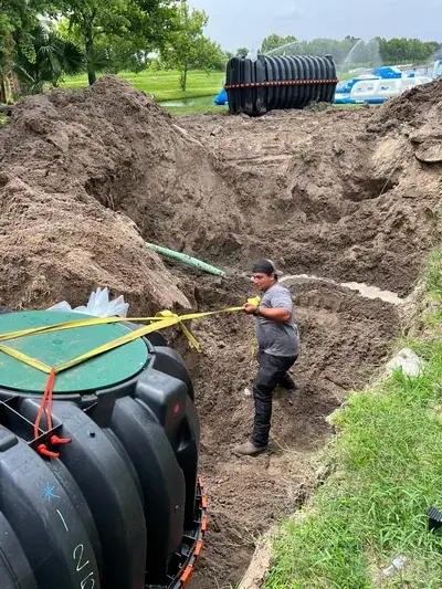 A man in a muddy dirt trench pulls a yellow strap attached to a large black tank with a green lid, while another similar tank and inflatable water park structures are visible in the background.