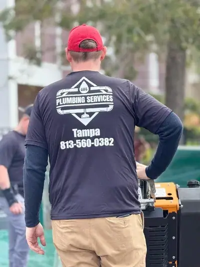 A person wearing a red cap and a black t-shirt with "CDS PLUMBING SERVICES Tampa 813-560-0382" printed on the back stands with their back to the camera. They are wearing khaki pants and are next to orange and black equipment.