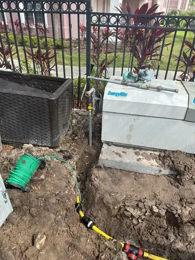 An outdoor scene showing a trench in the dirt with yellow gas lines and black connectors, a green coiled wire, and a grey utility box labeled "EnergyRite" next to a black storage container. A black metal fence and some foliage are visible in the background.