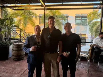 Three men stand smiling on a wooden deck, holding drinks. Palm trees and a building are visible in the background, suggesting an outdoor patio or rooftop setting.
