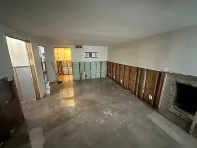 An interior room with a wet concrete floor and stripped-down walls, exposing studs and insulation. The room appears to be undergoing water damage remediation or renovation, with a fireplace opening visible on the right and a TV mount on the far wall.