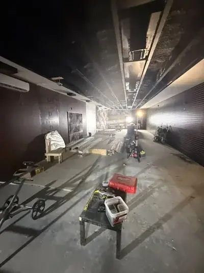 An indoor space under renovation with exposed ceiling beams, dark walls, and a concrete floor. Construction tools and materials, including a red toolbox and a container of screws, are visible on a table and scattered throughout the room.