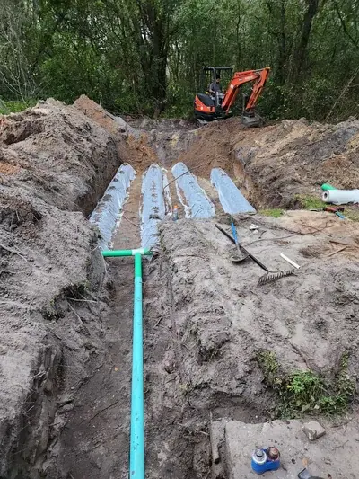 A construction site shows a large trench with several white septic drain field chambers and green PVC pipes installed. In the background, a person operates an orange Kubota mini-excavator on a dirt mound, surrounded by trees.