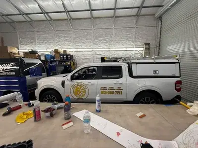 A white Ford Maverick pickup truck, branded for 'CDS SERVICE CO' with contact information, is parked inside a workshop. In the foreground, a table holds various items including bottles, tools, and a paper with children's drawings.