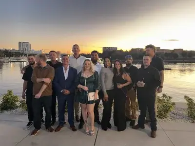 A group of about a dozen people stands outdoors by a body of water at sunset, with a city skyline and a "GO BOLTS" sign visible in the background.