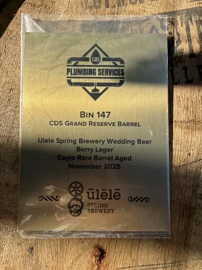 A golden plaque, partially covered by clear plastic, rests on a wooden surface. The plaque details a 'CDS Grand Reserve Barrel' containing 'Ulele Spring Brewery Wedding Beer Berry Lager,' 'Eagle Rare Barrel Aged,' with a release date of 'November 2025,' and features logos for CDS Plumbing Services and Ulele Spring Brewery.
