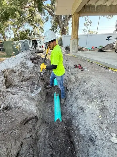 A construction worker in a hard hat and safety vest stands in a trench, using a shovel to move dirt around a green pipe. The surrounding area is a construction site with piles of dirt and a partially built structure.
