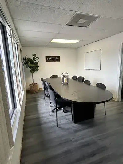 A conference room features a large dark oval table surrounded by black chairs, with a decorative lantern on top. Windows line the left wall, a potted plant sits in a basket, and a sign reading "AN ATTITUDE OF GRATITUDE" hangs on the back wall next to a whiteboard.