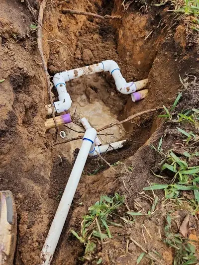 A trench dug in brown dirt reveals white PVC pipes, some connected with elbows and couplings, lying in muddy water. Tree roots are visible along the sides of the trench.