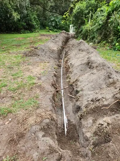 A long, narrow trench has been dug through a grassy area, with a white pipe laid along its bottom. Piles of excavated dirt line both sides of the trench, and dense green foliage forms the background.