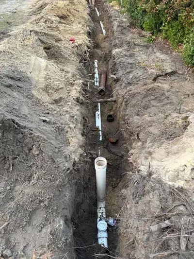 A long, narrow trench is dug into the dirt, revealing several white PVC pipes and one reddish-brown pipe laid along its length. Piles of excavated soil are visible on both sides of the trench, with some roots and debris scattered around.