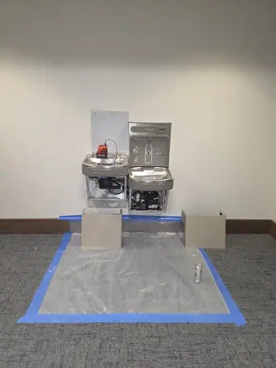 Two Elkay water bottle filling stations are mounted on a white wall, with their lower panels open revealing internal components. A red and black device sits on the left unit, and a plastic sheet is taped to the carpeted floor below, with two beige panels and a soda can on it.