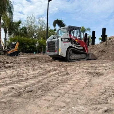 A Takeuchi TL8R2 compact track loader is parked on a dirt lot with tire tracks, next to a pile of excavated earth. Another piece of yellow construction equipment is visible in the background, with palm trees and other greenery under a cloudy sky.