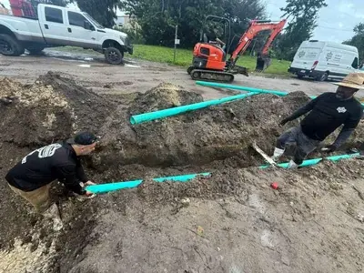 Two men are working in a muddy trench, installing green pipes. In the background, a mini excavator is visible, along with a white pickup truck and a white utility van.
