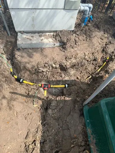 The photo shows yellow flexible pipes with black connectors laid in a dirt trench next to a large grey unit on a concrete slab, with grey pipes connecting to the unit. A green wire is also visible in the trench.