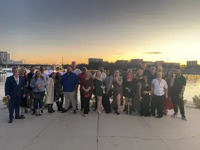 A large group of diverse adults stands outdoors on a paved area at sunset. In the background, a body of water reflects the orange sky, with city buildings, a 'GO BOLTS' sign, and a decorated boat visible.