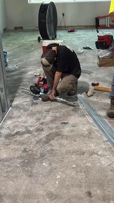 A worker wearing a cap and work clothes kneels on a concrete floor, using a red power saw to cut a metal channel. In the background, a large black industrial fan and various tools are visible in an unfinished room.