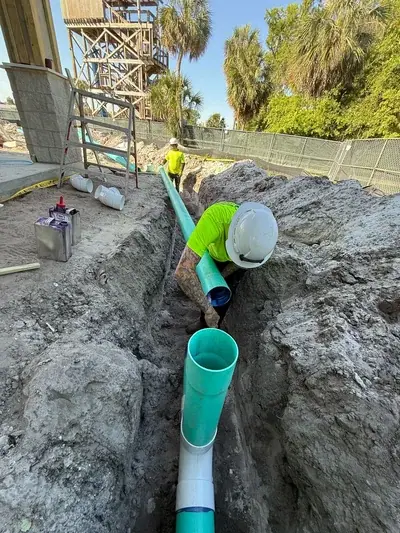 A construction worker in a neon shirt and hard hat is in a dirt trench, connecting a green pipe to another pipe. Another worker stands in the background near a large wooden structure, with palm trees and a chain-link fence visible under a clear sky.