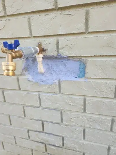 A brass outdoor water spigot with a blue handle is installed on a light yellow painted brick wall. A section of the brick around the spigot is cut out, revealing a light blue material and grey sealant.