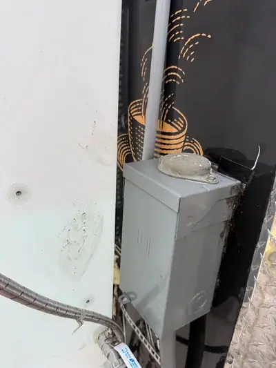 A close-up view of a grey electrical box with a conduit, mounted between a white wall and a black surface with orange patterns. A braided hose and valve are visible at the bottom left.