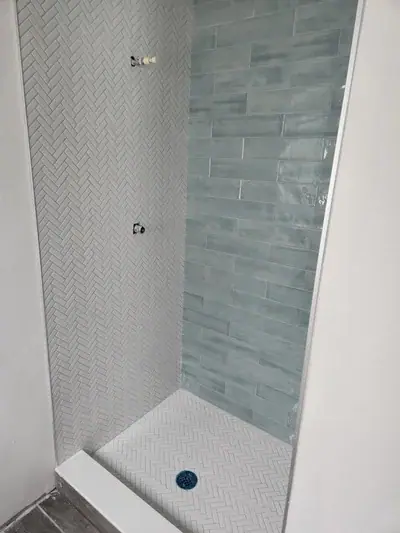 A shower stall under construction features light gray herringbone tiles on the left wall and floor, and blue-green rectangular tiles on the back wall. Exposed plumbing for a shower head and faucet are visible on the left wall, with a blue drain on the floor.