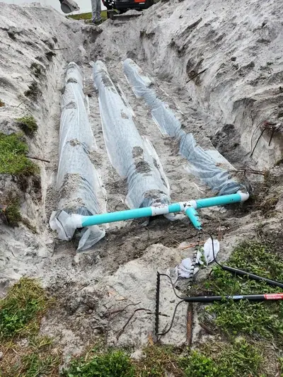 A trench dug in sandy soil contains three long, cylindrical objects wrapped in white fabric, partially covered with sand. A light blue PVC pipe assembly connects to one of the wrapped objects, with a person and an excavator visible at the top of the trench.