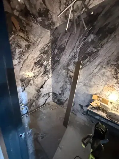 A room under construction with walls covered in black and white marble-patterned panels. The light gray floor shows some wetness, and a wooden board leans against the back wall, with tools and a lit lamp visible on a counter to the right.