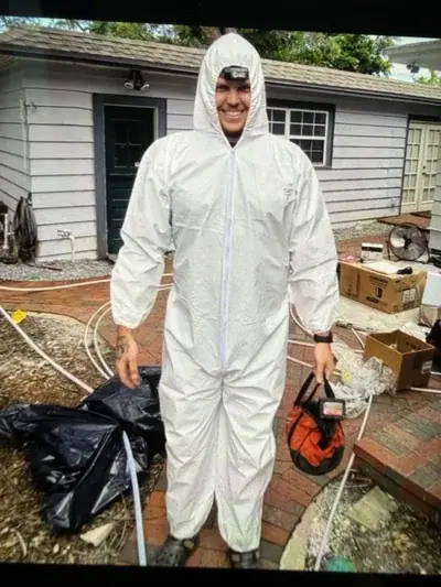 A man in a white hazmat suit with a headlamp smiles while standing on a brick patio, holding an orange and black bag. In the background is a house with light siding and various items like black trash bags, boxes, and a fan.