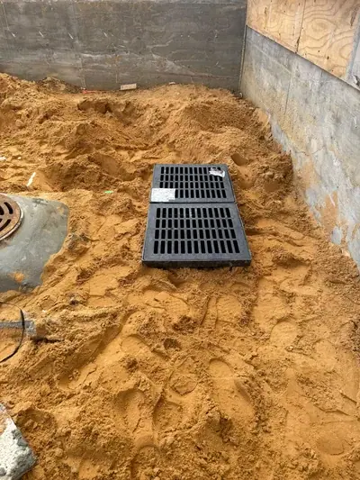 A construction site floor covered in a large pile of sand, featuring two black rectangular grates and a round metal manhole cover. Concrete walls and some plywood are visible in the background.