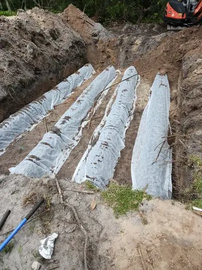 A deep trench in the earth contains several long, parallel objects covered in white fabric, likely part of a drainage or septic system installation. An orange excavator is visible in the background.