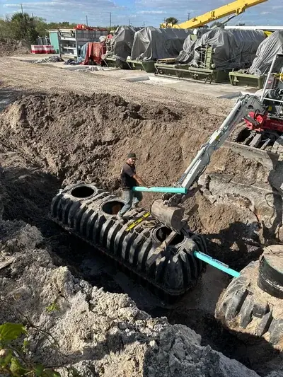 A worker stands on a large black corrugated tank in a deep trench, holding a light blue pipe. An excavator is positioned nearby, and another similar tank is partially visible to the right, connected by a pipe.