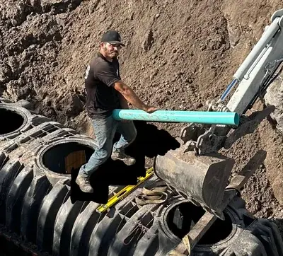 A plumber in a black shirt and cap stands on a large black ribbed tank in an excavated area, holding a light blue pipe. An excavator arm and a dirt wall are visible in the background.