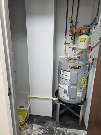 A utility room features a gray electric water heater on a metal stand, connected to an array of pipes, valves, an expansion tank, and a pump. To the left, a small utility sink with a yellow bucket is visible next to a white vertical column.